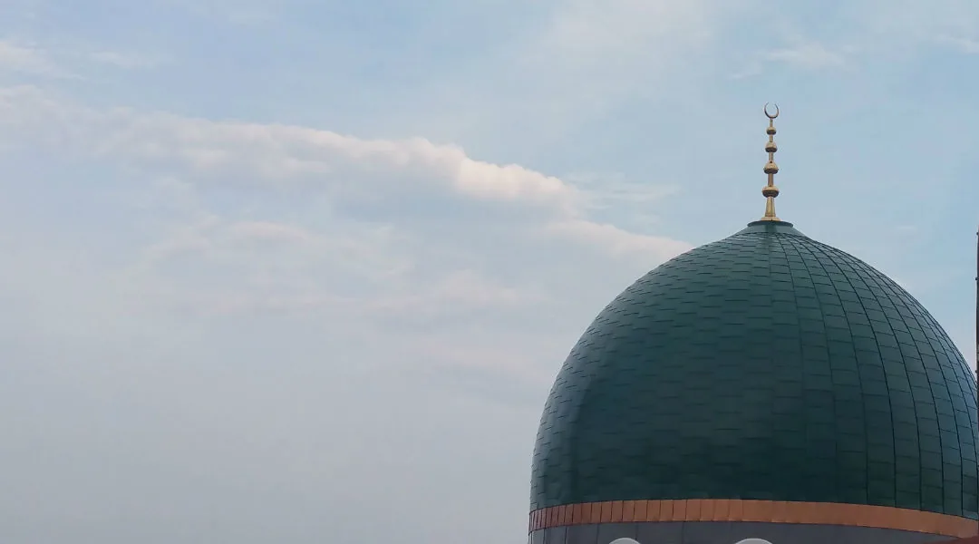 Green mosque dome with a crescent finial beneath a cloudy sky